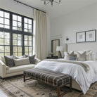 Elegant bedroom with beige curtains, large windows, plaid bench, and neutral bedding in a cozy setting.